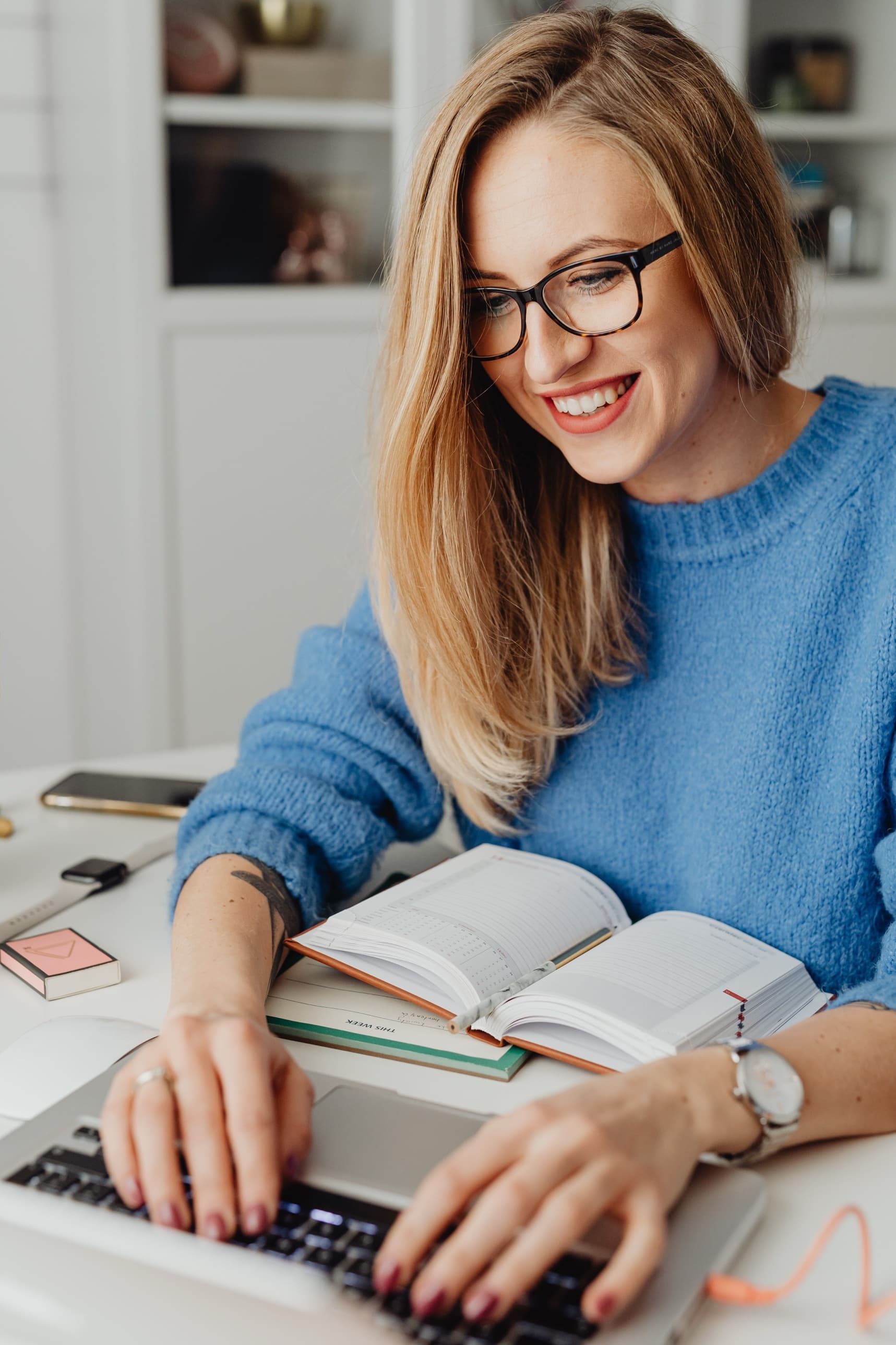A woman in a blue sweater at a desk, smiling at her laptop, an open planner beside her.